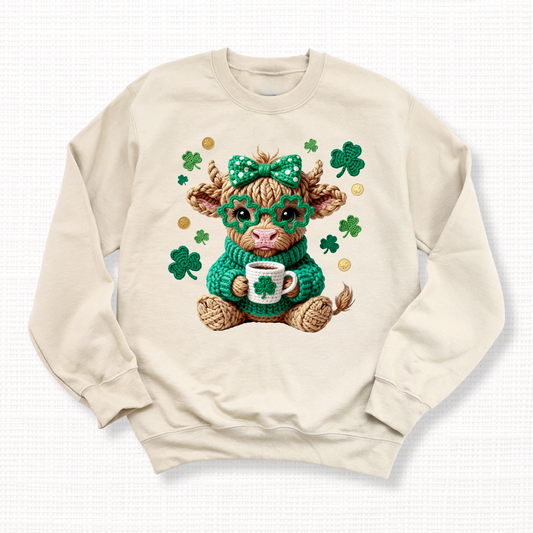 St. Patrick’s Day crochet Highland cow graphic sweatshirt with shamrocks, a green bow and glasses on a sand crewneck sweatshirt.