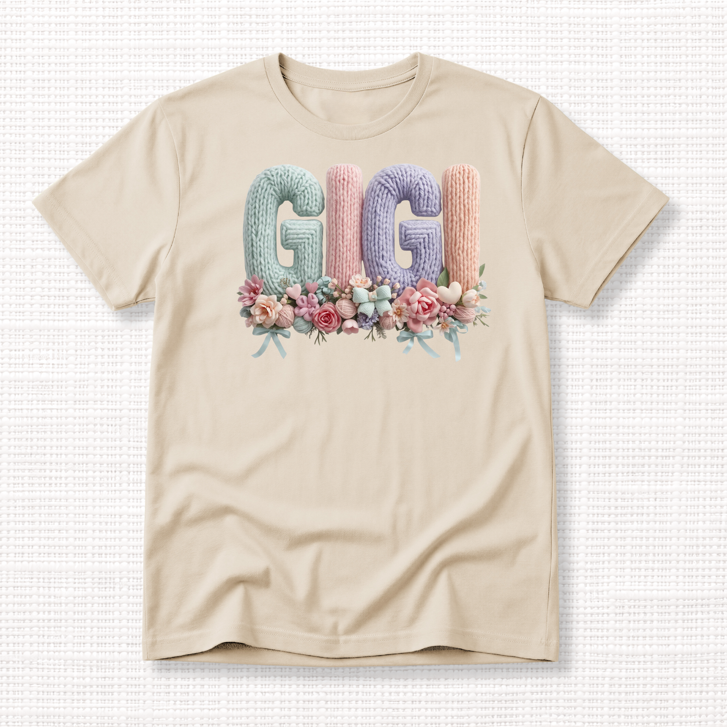 Beige t-shirt with “GIGI” in pastel crochet-style lettering, framed at the bottom with a bouquet of soft floral accents.