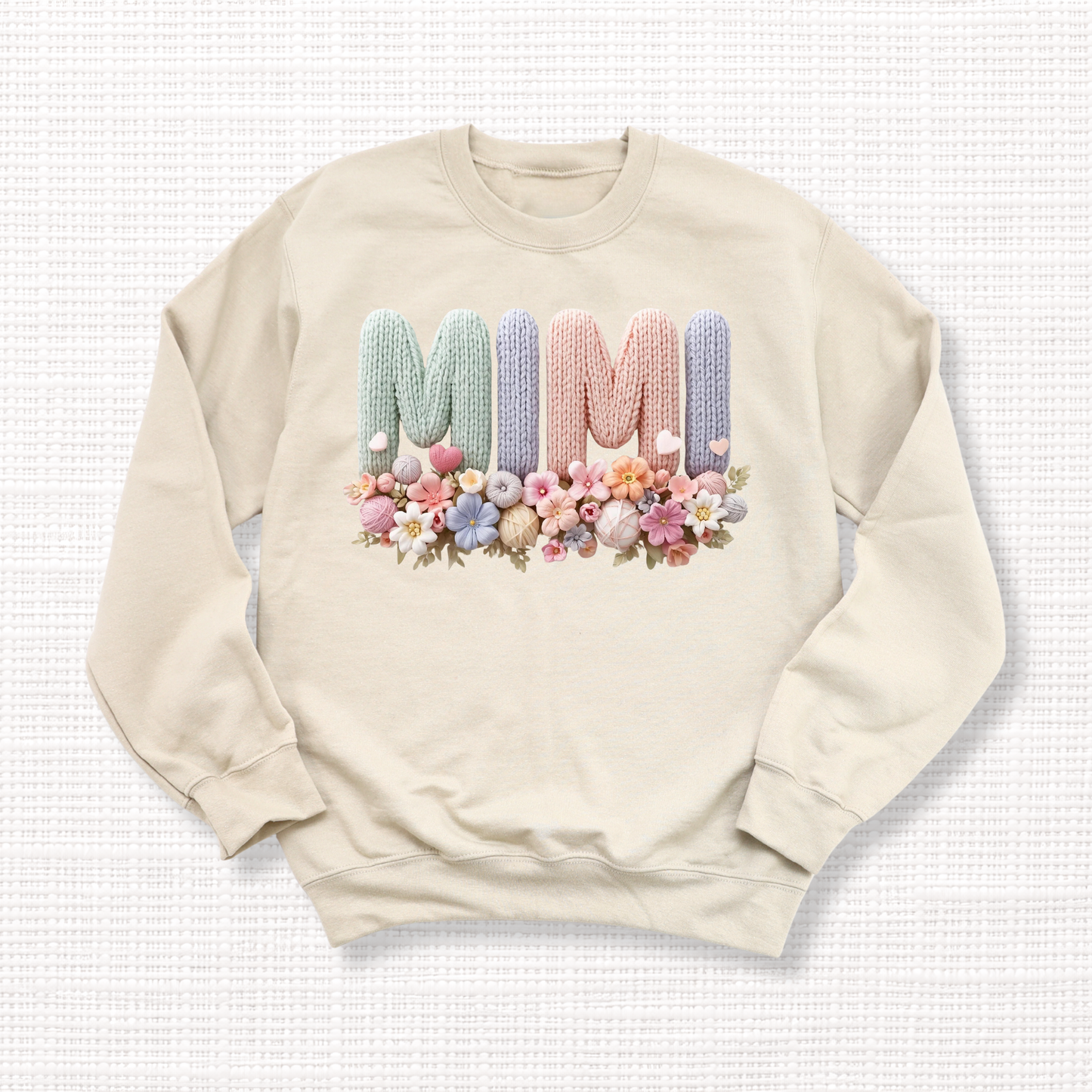 Beige crewneck sweatshirt with “MIMI” in pastel crochet-style lettering, framed at the bottom with a bouquet of soft floral accents.