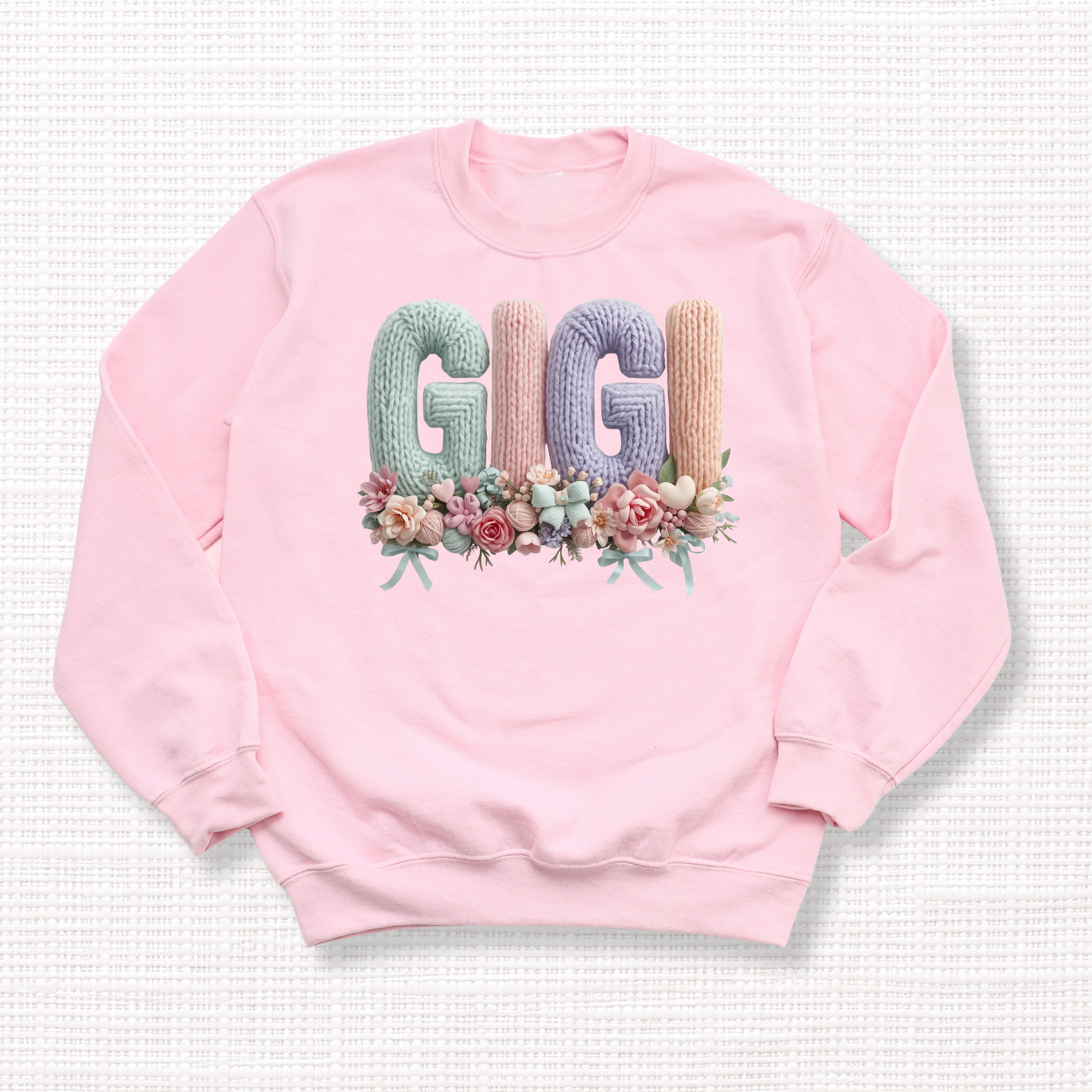 Light pink crewneck sweatshirt with “GIGI” in pastel crochet-style lettering, framed at the bottom with a bouquet of soft floral accents.
