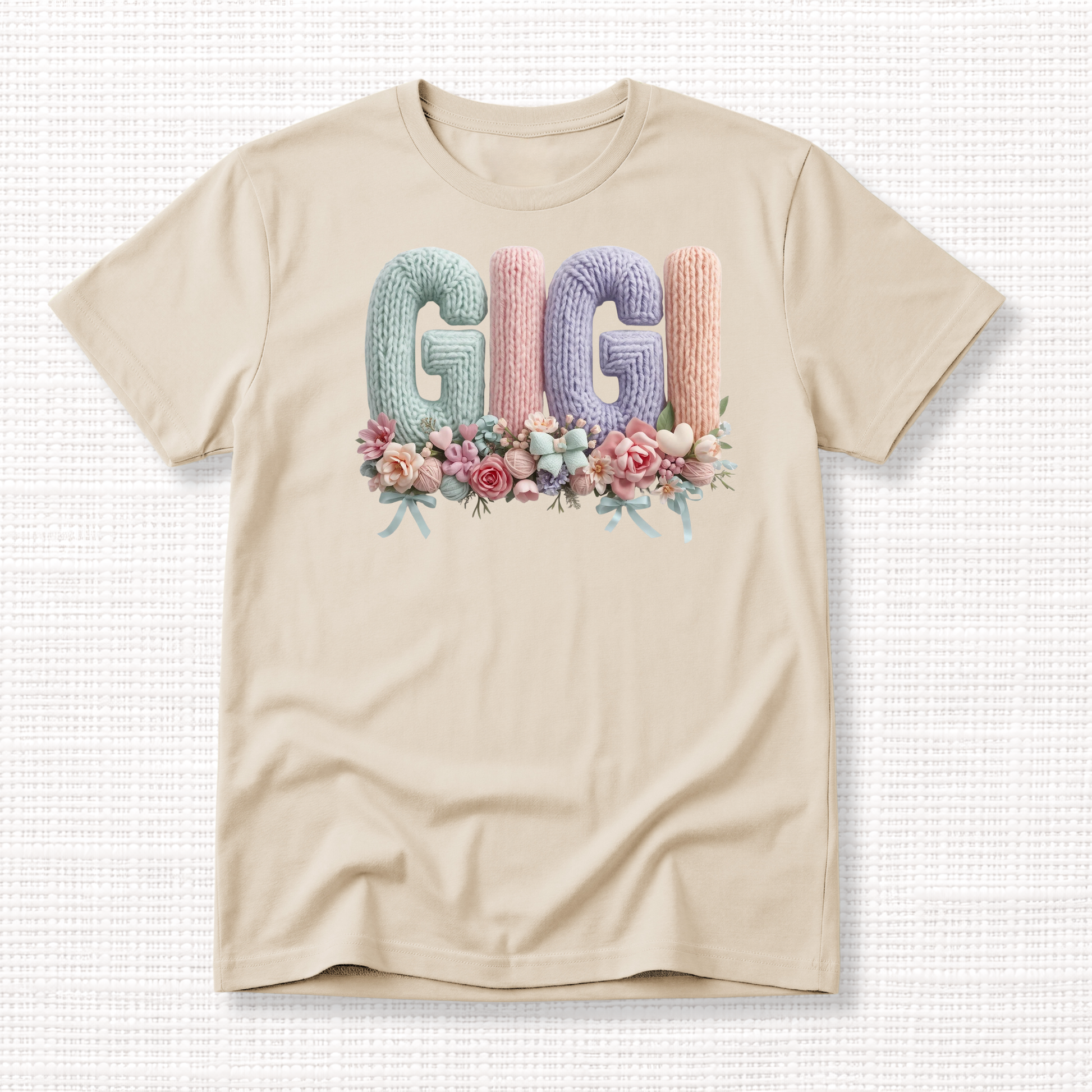 Beige t-shirt with “GIGI” in pastel crochet-style lettering, framed at the bottom with a bouquet of soft floral accents.
