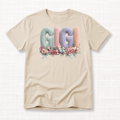 Beige t-shirt with “GIGI” in pastel crochet-style lettering, framed at the bottom with a bouquet of soft floral accents.
