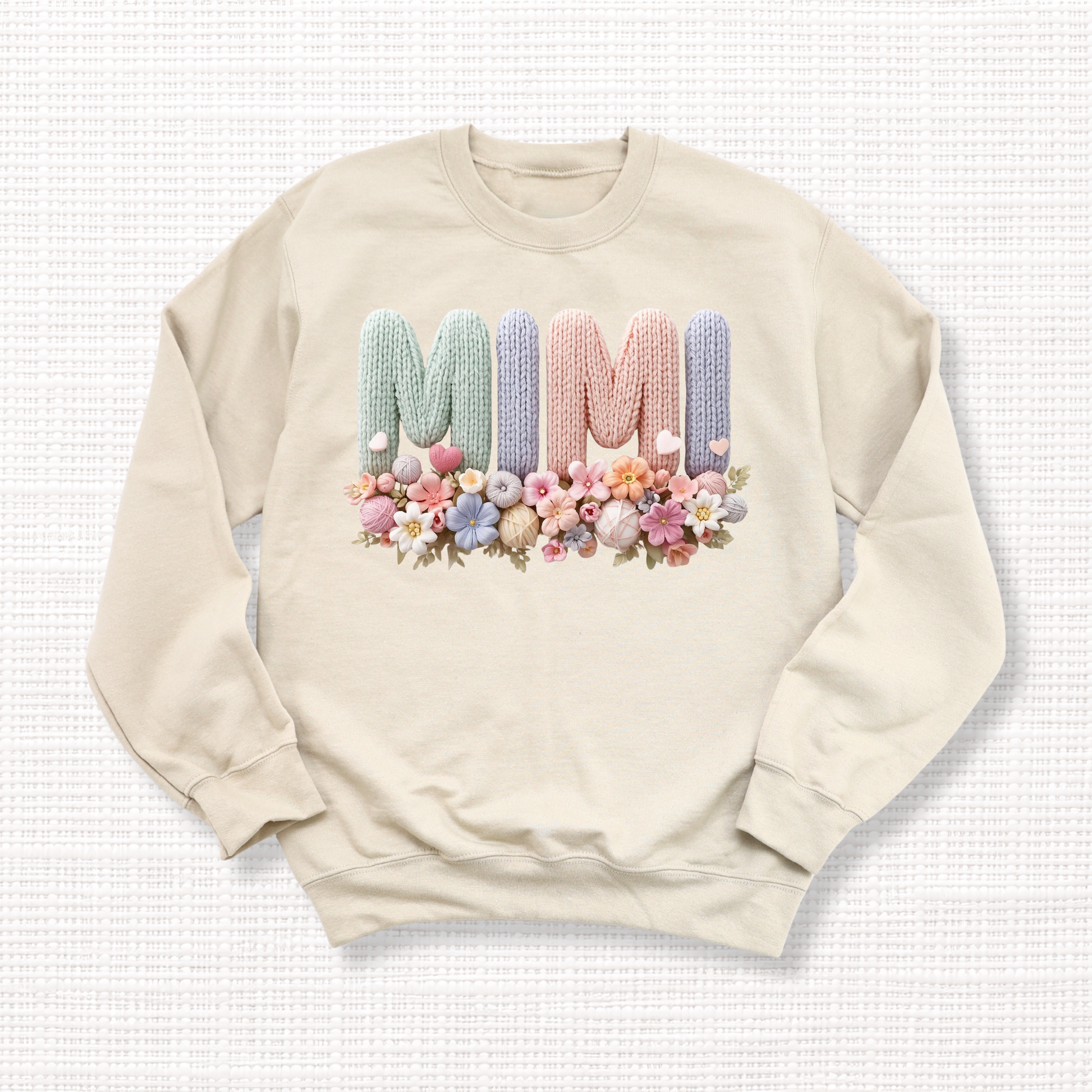 Beige crewneck sweatshirt with “MIMI” in pastel crochet-style lettering, framed at the bottom with a bouquet of soft floral accents.