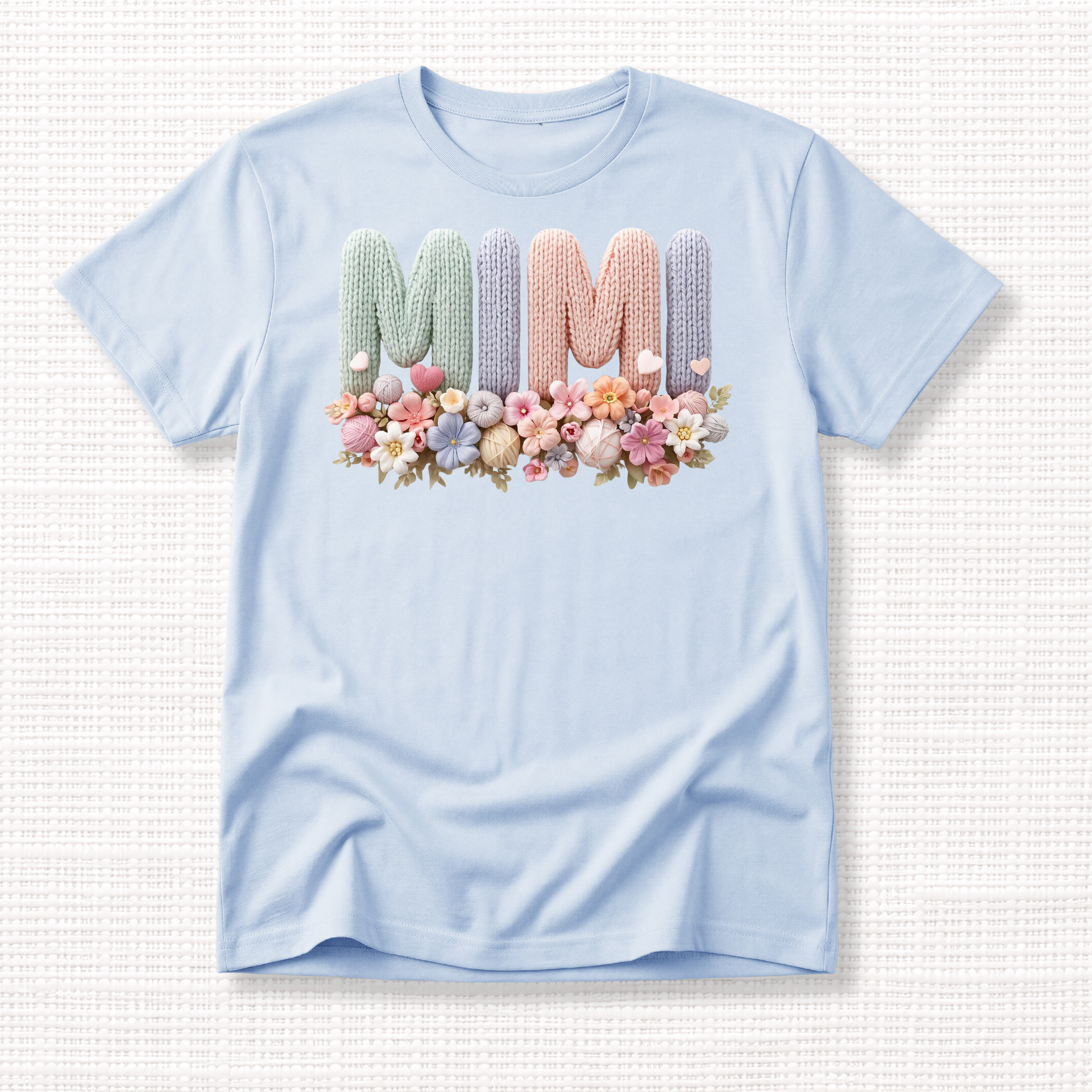 Light blue t-shirt with “MIMI” in pastel crochet-style lettering, framed at the bottom with a bouquet of soft floral accents.
