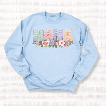 Light blue crewneck sweatshirt with “NANA” in pastel crochet-style lettering, framed at the bottom with a bouquet of soft floral accents.