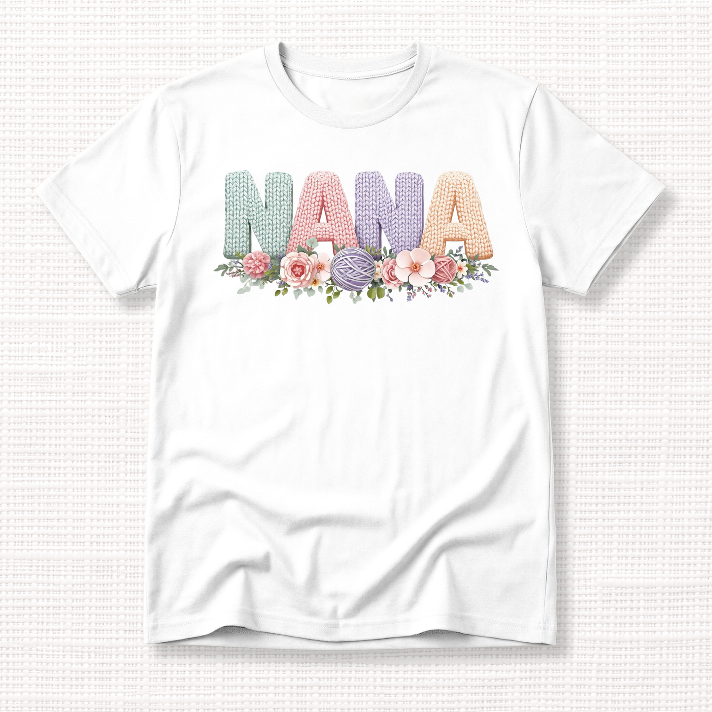 White t-shirt with “NANA” in pastel crochet-style lettering, framed at the bottom with a bouquet of soft floral accents.